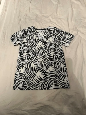 Svartvit t-shirt med bladprint Kappahl - Snygg t-shirt från Kappahl i storlek 146/152 med svartvitt bladmönster över hela plagget. Rund hals och korta ärmar. Materialet är mjuk bomull som känns skönt mot huden. Perfekt för dig som gillar grafiska prints och vill sticka ut lite extra.