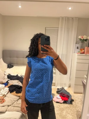 Blå blus med broderade hål och volang - Supersöt blå blus i bomull med broderade hål och volangdetaljer vid axlarna. Blusen har öppen rygg med knytband upptill och är ärmlös. Perfekt för varma dagar och ger en fräsch look. Passar både S, M jag har Xs