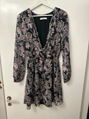 Blommig omlottklänning från MNG - Supersnygg kort omlottklänning från MNG med svart botten och blommigt mönster i rosa, beige och vitt. Klänningen har lång ärm, v-ringning och markerad midja. Perfekt för dig som gillar en feminin och trendig stil.