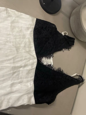 Svartvit linnetopp med spetsdetaljer - Säljer en snygg svartvit linnetopp med tunna axelband. Toppen har svart spets upptill och vit slät nederdel, vilket ger en elegant kontrast. Spetsen har fina fransiga kanter och toppen är ärmlös med v-ringad form. Perfekt till sommarens outfits!