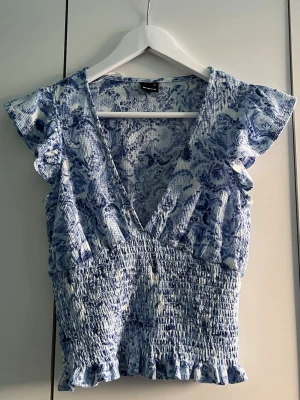Söt topp - Supersöt blus i blått och vitt. Modellen har v-ringning, volangdetaljer vid ärmar och nederkant samt smockad midja. 