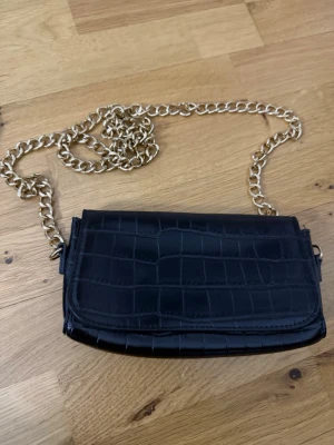 Svart axelväska med kedja - Snygg svart axelväska med krokodilpräglad yta och en rejäl guldfärgad kedja som axelrem. Väskan har ett stilrent och modernt utseende, perfekt för att lyfta din outfit. Materialet är skinnimitation och designen är enkel men trendig.