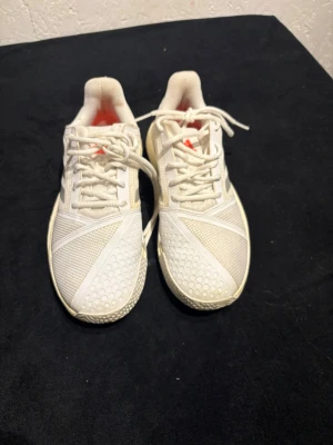 Vita Adidas sneakers med snörning - Säljer ett par vita Adidas sneakers med mesh och syntetdetaljer. Skorna har klassiska tre ränder på sidan, snörning och platt sula med mönstrad undersida. Insidan har en touch av orange och Bounce-dämpning för extra komfort. Perfekta för dig som gillar sportig stil.