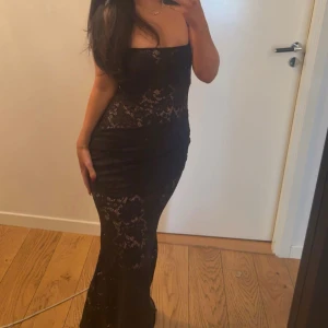 Spets klänning oh polly - Säljer en elegant svart långklänning med smala axelband och vacker spetsdetalj över hela klänningen. Klänningen har en figurnära passform och fyrkantig urringning som ger en stilren look. Genomskinlig 