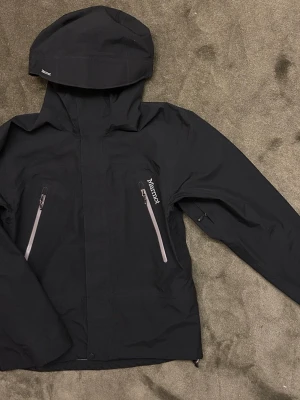 Svart Marmot vindjacka med huva - Marmot Gore‑Tex Performance Shell, strl S skaljacka i svart. Gore‑Tex. Jackan är sparsamt använd, hr dock ett litet hål på ärmen som är lagat. 