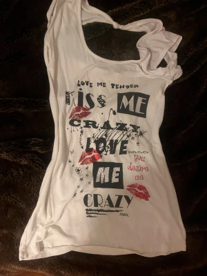 Vitt y2k linne  - Säljer en vit topp med coolt tryck i svart och rött med text och läppar framtill. Toppen har en unik knutdetalj vid halsen och är ärmlös med rundad , perfekt för att sticka ut med ett statement-plagg. Jag köpte linnet seckonhand och den har ingen lapp så vet inte storlek eller var den kmr ifrån men ja skulle säga S 🫶🏼