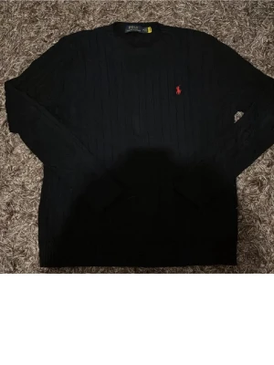 Svart stickad tröja från Polo Ralph Lauren - Svart stickad tröja från Polo Ralph Lauren med diskret ribbad struktur och klassisk röd logobrodyr på bröstet. Tröjan har långärmad design och rund hals, perfekt för en stilren och enkel look.