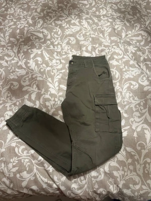 Olivgröna cargopants från Lager 157 - Säljer ett par olivgröna cargopants från Lager 157 med elastiska muddar vid bensluten och stora sidofickor. Byxorna har normal passform och är tillverkade i bomull, perfekta för en chill och avslappnad stil.