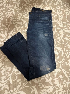 Slim fit jeans från Jack & Jones - Mörkblå slim fit jeans från Jack & Jones, modell Tim. Snygga slitningar på benen och klassisk femficksdesign. Jeansen är tillverkade i bomull med en rak passform och har en knappstängning i midjan. Perfekt för dig som gillar en smal siluett.