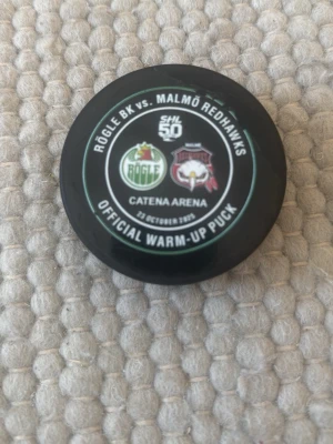 Warm upp puck  - Bilderna visar en officiell hockeypuck från matchen Rögle  vs. Malmö Redhawks i Catena Arena. Pucken är svart och rund med tryck på ovansidan.