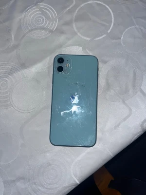 iPhone 11 - Säljer en blå iPhone 11. Baksidan har tydliga sprickor och repor, samt viss slitage på kanterna. Skärmen har fingeravtryck men inga synliga sprickor. Dubbelkamera och klassisk Apple-design. Perfekt för dig som vill ha en prisvärd iPhone trots kosmetiska defekter.