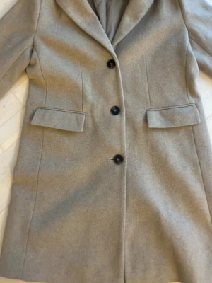 Beige kappa från Ellos med knappar - Snygg beige kappa från Ellos med klassisk krage och tre  knappar framtill. Jackan har två fickor med lock och är helfodrad på insidan. Perfekt för dig som gillar stilrena och tidlösa plagg.