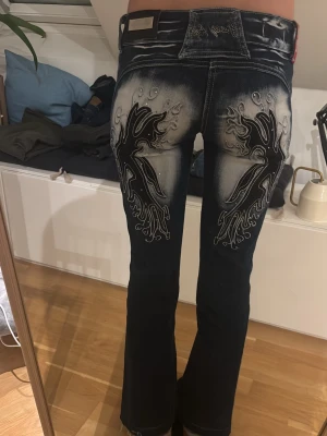 Young age jeans med broderad design - Säljer ett par mörkblå Young age jeans med broderad design och pärldetaljer på bakfickorna. Jeansen har bootcut-snitt och låg midja.