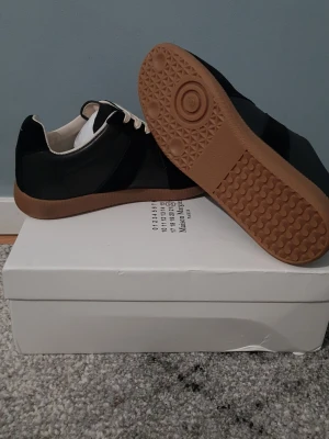 Maison Margiela svarta sneakers - Maison Margiela sneakers i svart med kontrasterande vita snören och gummisula i brunt. Skorna har en klassisk rund tå och platt sula med unikt mönster undertill. Materialet är en mix av skinn och mocka, vilket ger en lyxig känsla och snygg look.