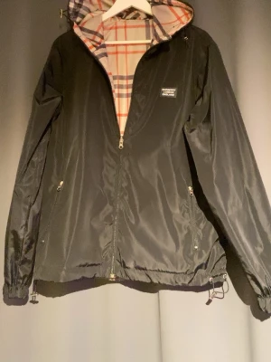 Burberry vindjacka med vändbar design - Snygg vändbar vindjacka från Burberry med klassiskt rutigt mönster på ena sidan och svart med logotyp på andra. Jackan har huva, dragkedja med Burberry-detalj, två fickor och justerbar nederkant. Perfekt för dig som vill ha både stil och funktion.