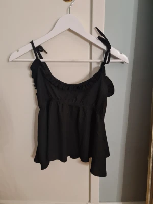 Svart linnetopp från 157 XS - Säljer en svart linnetopp från 157 i storlek XS. Toppen har tunna axelband med knyt och volangdetaljer längs halsringningen. Materialet är lätt och har en skrynklig struktur som ger en cool look. Perfekt för varma dagar och enkel att matcha med jeans eller kjol.