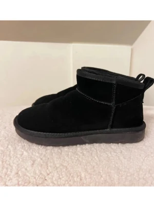 Svarta mockaboots med platt sula - Mysiga svarta boots i mocka med platt sula och rund tå. Skorna har en enkel design med låg skaft och dragflik bak, perfekta för kalla dagar. Passar till både jeans och mjukisbyxor.