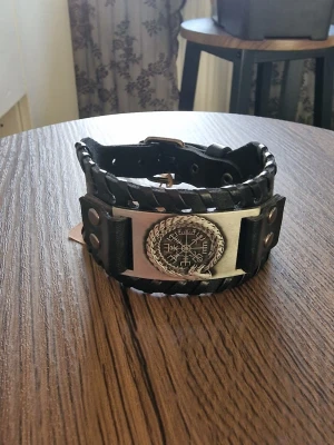 Armband vintage nordisk vikingkompass - Unikt svart PU läderarmband med flätade kanter och en stor metallplatta framtill. Plattan har ett vikingainspirerat mönster och är dekorerad med nitar. Justerbar längd med spänne. 27,4 cm långt.  Perfekt accessoar för dig som gillar en tuff och alternativ stil.