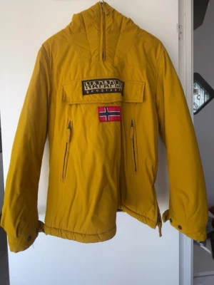 Gul vindjacka från Napapijri - Säljer en gul vindjacka från Napapijri med huva och cool norsk flagga på bröstet. Jackan har dragkedjor på fickorna och en stor ficka framtill med logotyp. Perfekt för dig som vill sticka ut med färg och stil.