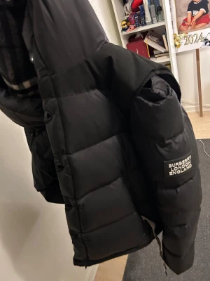 pufferjacka/väst från Burberry  - Säljer en svart pufferjacka från Burberry London England i storlek M. Jackan har en klassisk pufferdesign med stora vadderade paneler, dragkedja och knappar framtill samt en huva fodrad med mönstrat tyg. Den görs även till en väst, då det går att ta av och sätta på armarna.