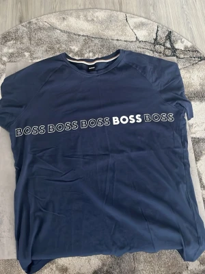 Mörkblå t-shirt från BOSS - Snygg mörkblå t-shirt från BOSS med vit logotyptext tryckt flera gånger över bröstet. Rund halsringning med en diskret randig detalj på insidan av kragen. Perfekt för dig som gillar stilrena och moderna plagg. Helt ny, 10/10 skick!