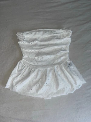 Vit bandeau tubtopp med broderade detaljer - Säljer en vit bandeau tubtopp med broderade hålmönster och volang nedtill. Toppen är axelbandslös och har en söt, feminin look med scallop-kant. Perfekt för varma dagar och sommarhäng.