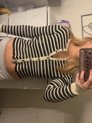 Randig croppad kofta med knappar - Säljer en svartvit randig croppad kofta med långa ärmar och knappar framtill. Tröjan har ribbade muddar och en tight passform som framhäver midjan. Perfekt för dig som gillar en trendig och stilren look.(de är inte fläck på tröjan de är spegeln)