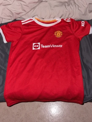 Manchester United Ronaldo 7 tröja - Säljer en röd Manchester United matchtröja med vita detaljer och tryck. Framsidan har TeamViewer-logga och klubbmärke, baksidan har Ronaldo och nummer 7 i vitt. Perfekt för fotbollsfans som vill visa sitt stöd för laget.