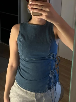 Blå ärmlös denim topp - Säljer en blå topp i denim med ärmlös design och snörning på sidan. Toppen har en figurnära passform och dragkedja längs ryggen för enkel på- och avtagning.