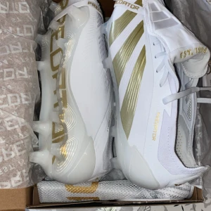 Adidas Predator fotbollsskor vit/guld - Säljer ett helt nya par Adidas Predator Elite FG  fotbollsskor i vitt med snygga guldfärgade detaljer och ränder. Skorna har snörning och platt sula med dobbar för gräsplan. Materialet är syntet och designen är riktigt clean och modern, perfekt för dig som vill sticka ut på planen. Skorna har använts bara en träning, säljs eftersom de är mindre i storlek på mig. Säljs med allt packningen. 