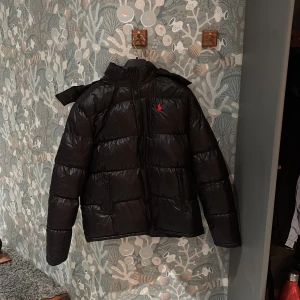 Svart dunjacka  - Snygg svart dunjacka med en glansig finish, huvaoch dragkedja framtill. Perfekt för kalla dagar och perfekt till hösten och vintern. Priset är inte hugget i sten.