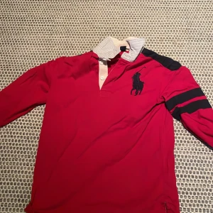 Röd pikétröja från Polo Ralph Lauren - Röd långärmad pikétröja från Polo Ralph Lauren med klassisk krage i blåvitrandigt tyg och stora svarta loggan på bröstet. Svarta ränder på ena ärmen och knappar vid halsen. Perfekt för en sportig och stilren look.