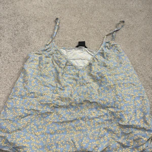 Blommig blå linnetopp från Lindex - Supersöt blå linnetopp från Lindex med tunna axelband och v-ringning. Toppen har ett gult blommigt mönster över hela och är perfekt för varma dagar. Lätt och luftig känsla, passar till jeans eller kjol.