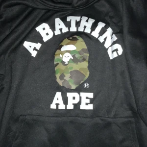 Svart BAPE hoodie med camo-logo - Svart hoodie från A Bathing Ape med klassisk camo-apa på bröstet och stor vit text. Tröjan har huva med snörning och känguruficka framtill. Ribbade muddar och ikonisk BAPE-tag på ärmen. Perfekt för dig som gillar streetwear.