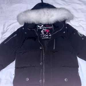 Svart Moose Knuckles dunjacka med pälskrage - Svart dunjacka från Moose Knuckles med fluffig vit pälskant på huvan. Jackan har flera dragkedjor, tryckknappar och en broderad patch på insidan. Perfekt för kalla dagar med sin tjocka vaddering och snygga detaljer.