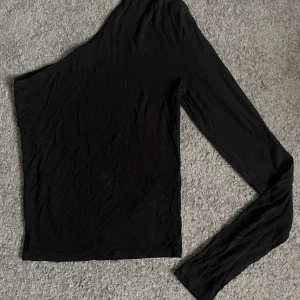 Svart oneshoulder topp - Snygg svart oneshoulder topp med lång ärm på ena sidan. Toppens minimalistiska design gör den lätt att matcha med olika outfits. Tillverkad i mjukt material som sitter skönt mot kroppen.