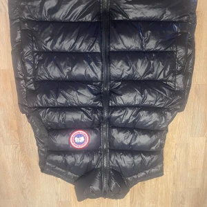 Canada Goose väst - Snygg svart dunväst från Canada Goose med klassisk logga på bröstet. Västen har quiltad design, dragkedja framtill och en stilren look som funkar till många outfits. Perfekt för kyliga dagar när du vill hålla värmen utan att tumma på stilen.