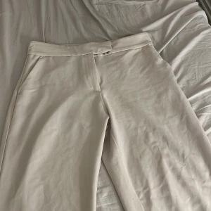 Beiga kostymbyxor med rak passform - Snygga beiga kostymbyxor med rak siluett och hög midja. Byxorna har diskret knappstängning och är tillverkade i ett mjukt bomullsmaterial. Perfekt för en clean och stilren look. Passar dig som gillar minimalistisk stil. står inte stolen eller vart de är ifrån men skulle gissa på bikbok och storlek M 