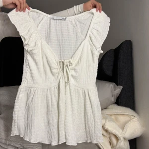 Vit blus med volang och knyt - Supersöt vit blus från 157 med volangdetaljer på axlarna och v-ringning med knyt framtill. Blusen har en lätt rynkad struktur och är tillverkad i ett mjukt, luftigt bomullsmaterial. Perfekt för dig som gillar romantisk och feminin stil.