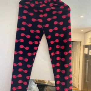 Svarta leggings med rosa rosett-mönster - Säljer ett par svarta leggings med elastisk midja och ett coolt rosa rosett-mönster över hela byxan. Perfekta för dig som vill sticka ut med färg och mönster. Sköna och stretchiga, passar till många olika outfits.