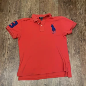 Röd pikétröja från Polo Ralph Lauren - Storlek 14-16 L junior. Snygg röd pikétröja från Polo Ralph Lauren med blått broderat logomärke på bröstet och en blå siffra på ärmen. 