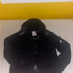 Svart Moncler pufferjacka montgenevre - Svart pufferjacka från Moncler med huva och klassisk logga på ärmen. Jackan har dragkedja framtill, fickor med lock och en blank insida med serietidningsmärke. Perfekt för kalla dagar och har en riktigt schysst streetstil.  I bra condition också