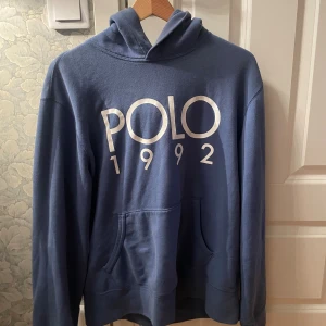 Blå hoodie från Polo Ralph Lauren - Snygg blå hoodie från Polo Ralph Lauren med stor vit logga och årtalet 1992 tryckt på bröstet. Tröjan har huva, känguruficka och långa ärmar. Perfekt för dig som gillar klassisk streetstyle och vill ha en bekväm och stilren look.