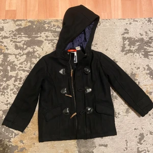 Svart Barn duffeljacka från Okaou  - Svart duffeljacka från Okaou med huva och klassiska träknappar. Jackan har quiltat blått innerfoder och två stora fickor framtill. Perfekt för kyliga dagar och har en snygg, tidlös look med dragkedja och knäppning. Jackan är till barn, storlek 116 barn. Pris kan diskuteras! Endast seriösa köpare, tack! ☺️ 