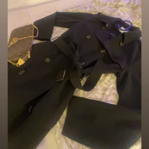 Svart trenchcoat med bälte - Snygg svart trenchcoat med klassisk krage, dubbelknäppning och bälte i midjan. Jackan har långa ärmar och stilrena knappar framtill. Perfekt för dig som vill ha en clean och tidlös look under höst och vår.