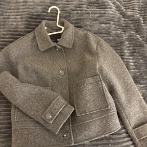 Grå overshirt med stora fickor - Snygg grå overshirt  med klassisk krage, stora framfickor och knappar framtill. Jackan har en boxig och lite croppad passform, perfekt att slänga över en hoodie eller t-shirt. Materialet är ullblandat och känns mjukt och varmt.