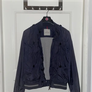 Moncler Bomber-/Militärjacka Mörkblå - Säljer denna stilrena Moncler jackan eftersom den inte passar mig längre. Skicket är 8/10 enda märkvärdiga defekten är lite slitage på vänstra armbågen. Nypris 10 000kr. Modellen heter ”DELONIX GIUBBOTTO DELONIX JACKET”. Köpt från en kompis, så har inget kvitto, men den är äkthetsverifierad genom Monclers hemsida. 