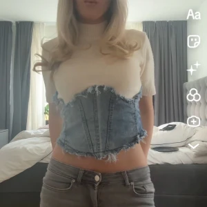 Vit stickad topp med denimkorsett - Unik topp med kort ärm i vit stickad och fastsydd denimkorsett över. Korsetten är i ljusblå jeans med råa, fransiga kanter och markerade sömmar. Hög halsringning och croppad passform.