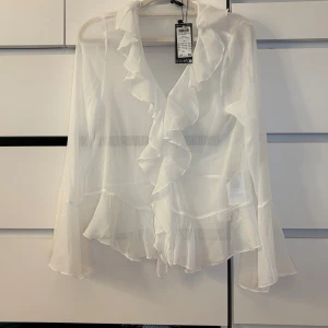 Vit volangblus med v-ringning - Säljer en vit, transparent blus från Gina Tricot med v-ringning och volangdetaljer längs halsen och nederkanten. Blusen har långa, lätt utsvängda ärmar och ett flowigt, romantiskt fall. Perfekt för dig som gillar en feminin och trendig stil.