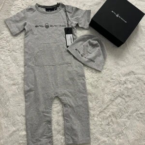 Sail Racing babyset i presentbox 6–12 mån  - Sail Racing babyset, jumpsuit och mössa i grått. Setet innehåller en mjuk kortärmad body med ficka framtill och tryckknappar längs benen, samt en matchande mössa med loggan. Storlek 6–12 månader 100% bomull 🌟HELT NY med tag kvar! 🌟 Kommer i original presentförpackning. perfekt som present! 🩶